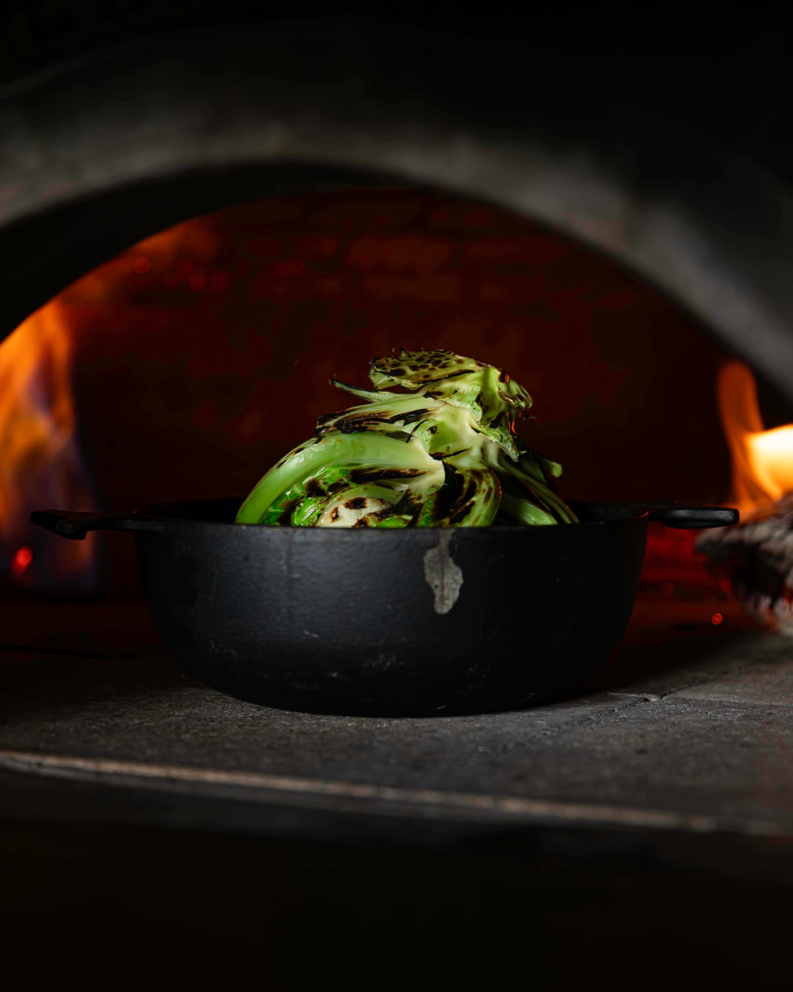 Charred greens in a cast-iron pot pulled from a wood-fired oven, embers glowing in the background