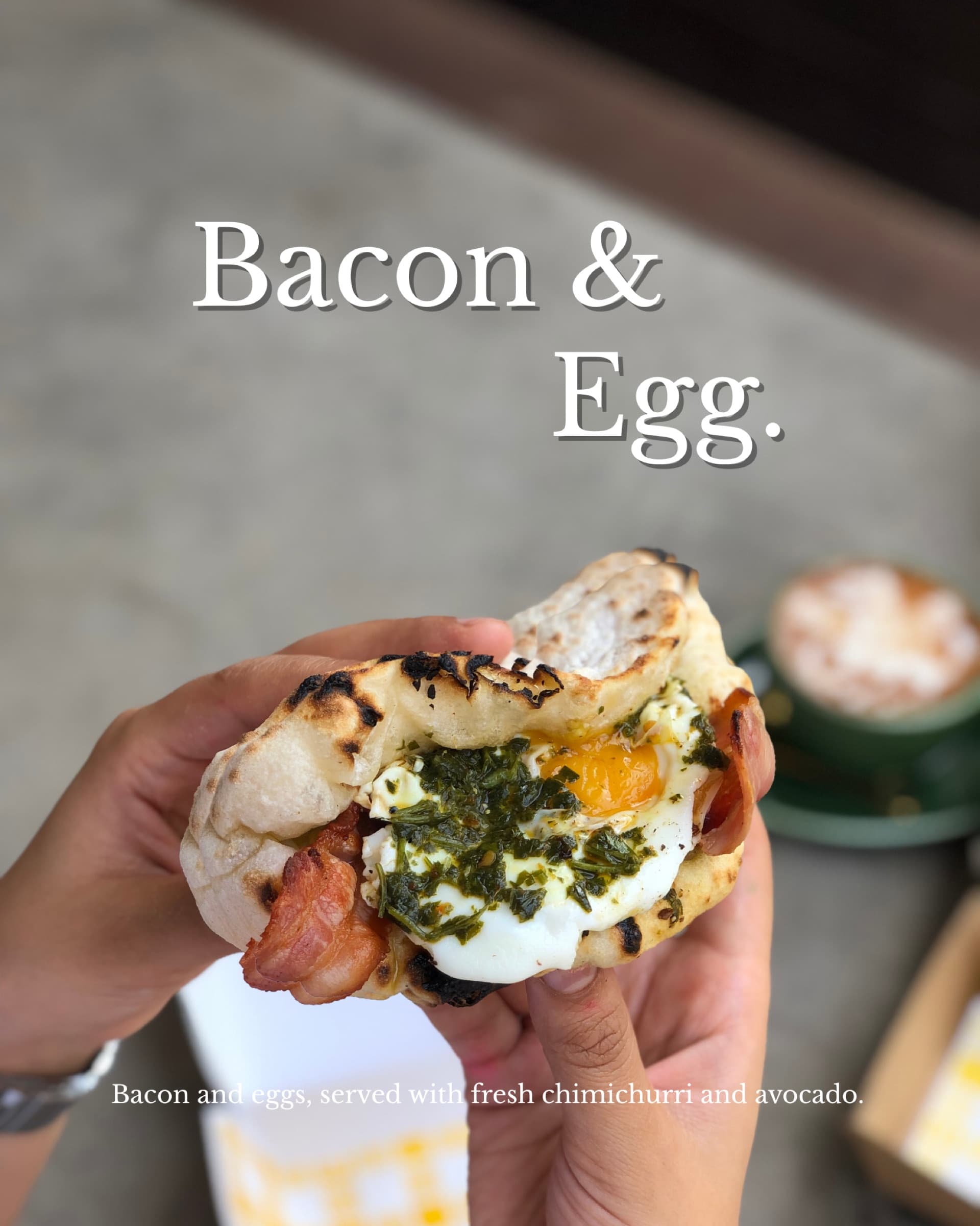 Bacon & Egg breakfast ozzo held in hand — bacon, eggs, chimichurri and avocado, with a coffee in the background