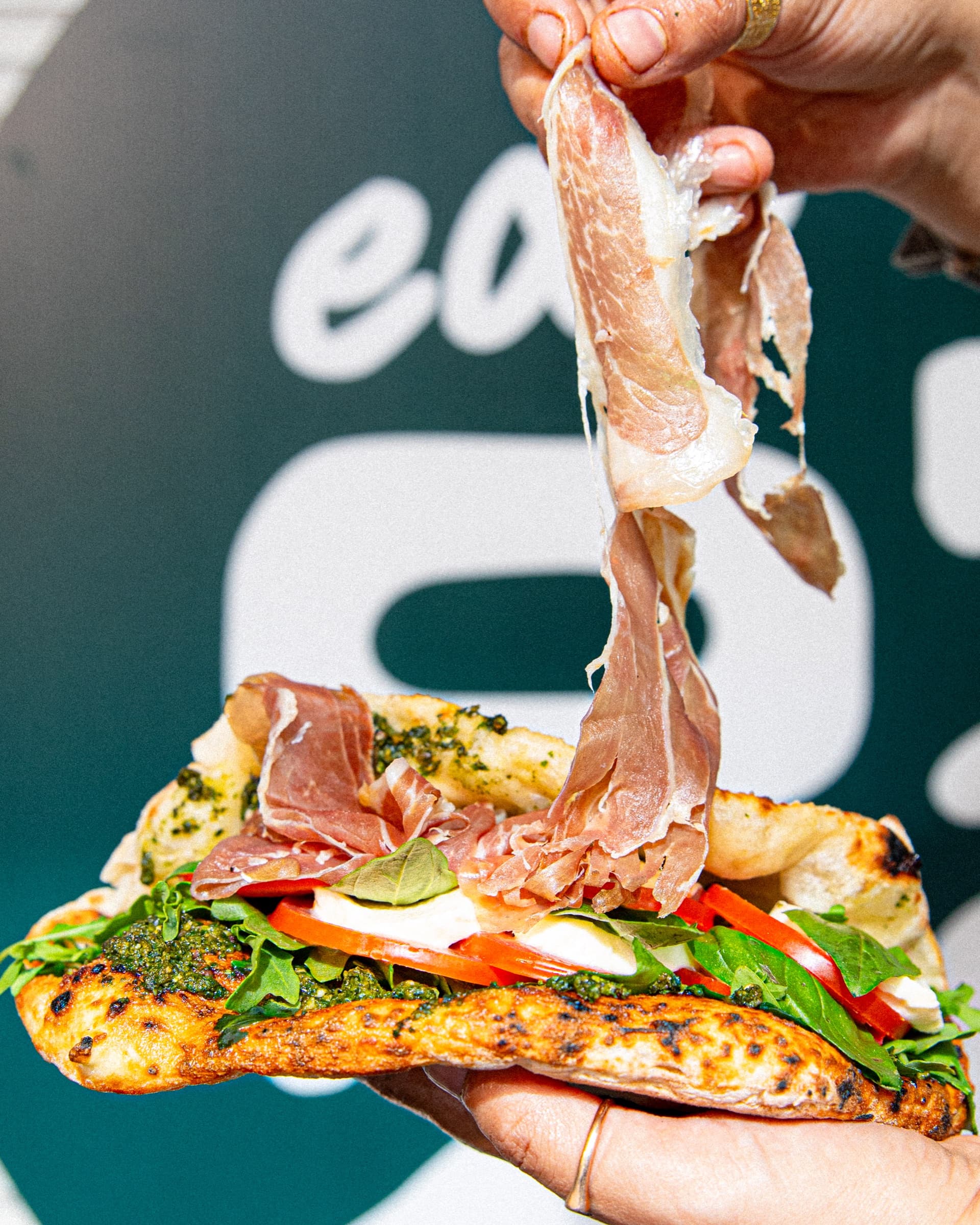 An open ozzo lifted in front of the Marrickville ozzo signage — prosciutto, bocconcini, basil pesto, tomato, rocket