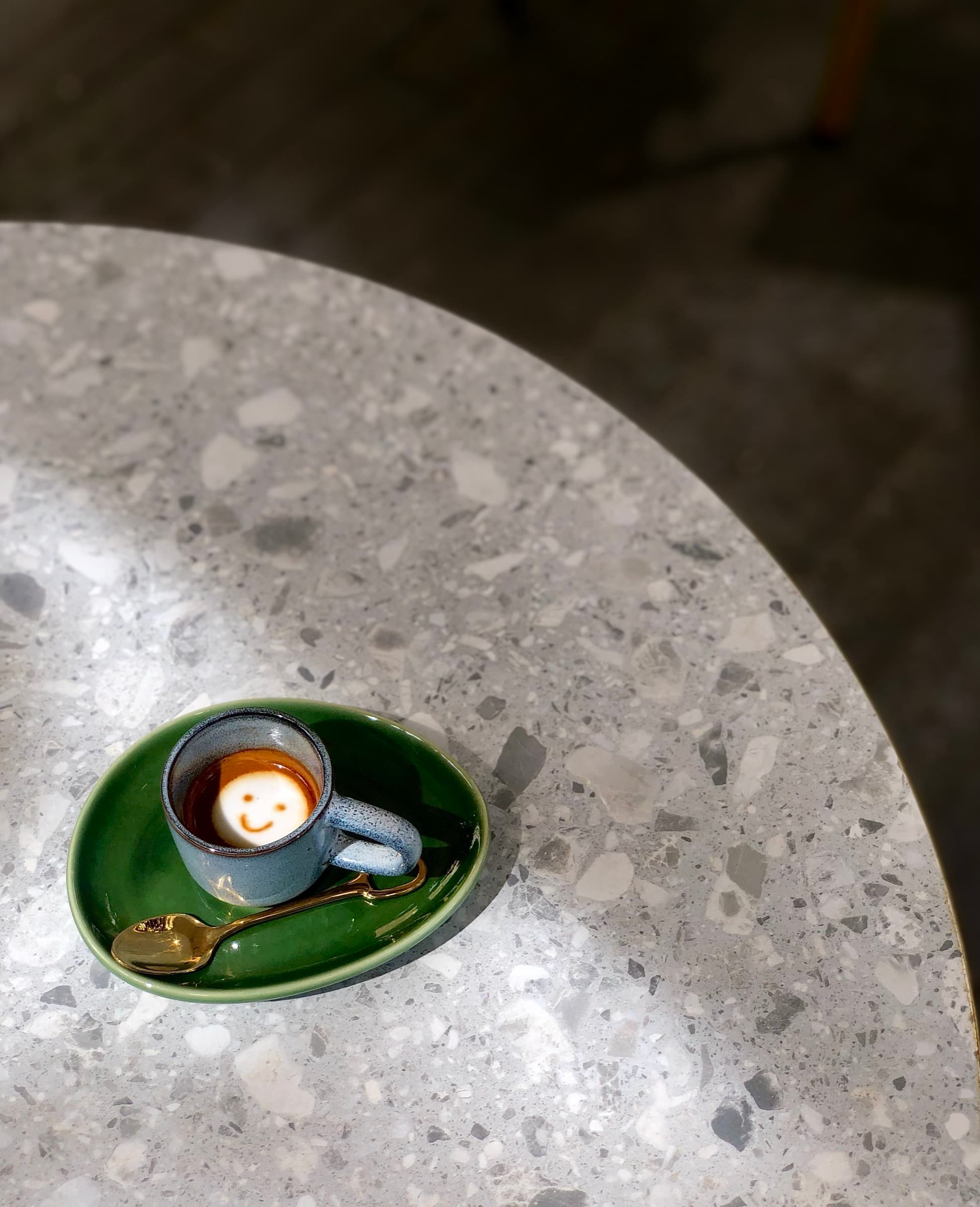 A single-origin espresso shot with a tiny smiley-face latte art rosetta, on a green saucer over terrazzo