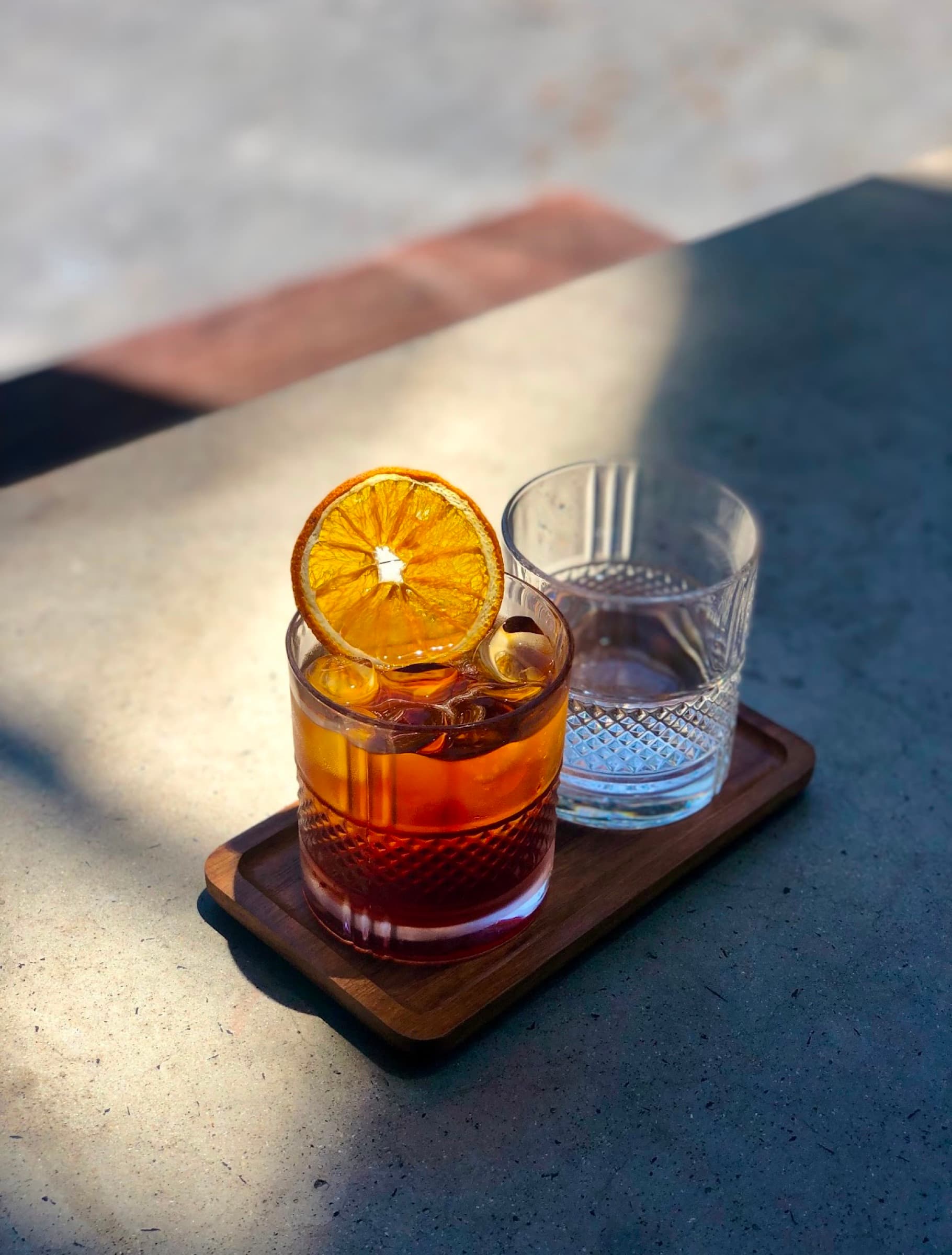 A Negroni with a dried orange wheel on a wooden serving board, water carafe beside it
